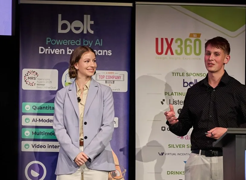 Kate Besel and Drake on stage at UX360 North America, in front of the conference banners: Drake, in a black shirt beside the lectern, mid-explanation; Kate, in a lilac blazer, smiling.